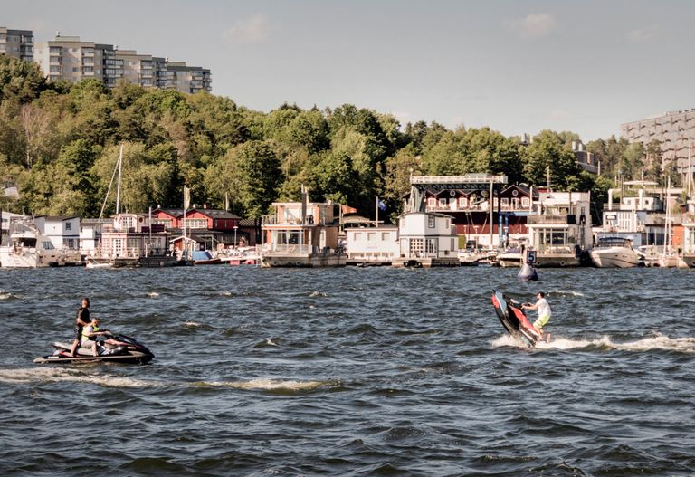 Två personer åker vattenskoter på vågigt vatten med en bakgrund av färgglada byggnader, träd och en klar himmel.