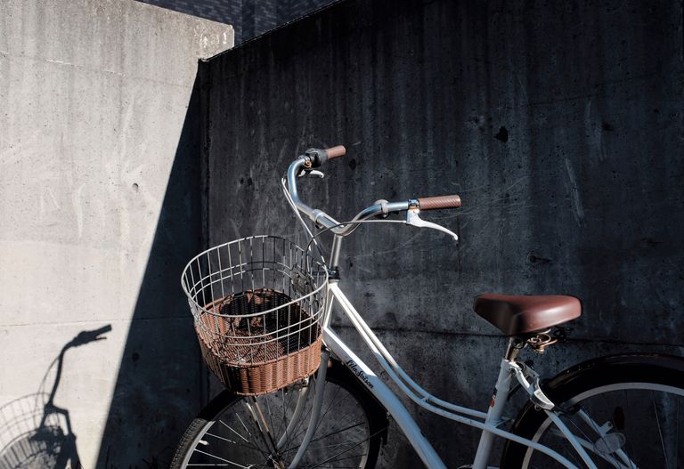 En vit cykel med trådkorg och brun sits står parkerad mot en mörk betongvägg.