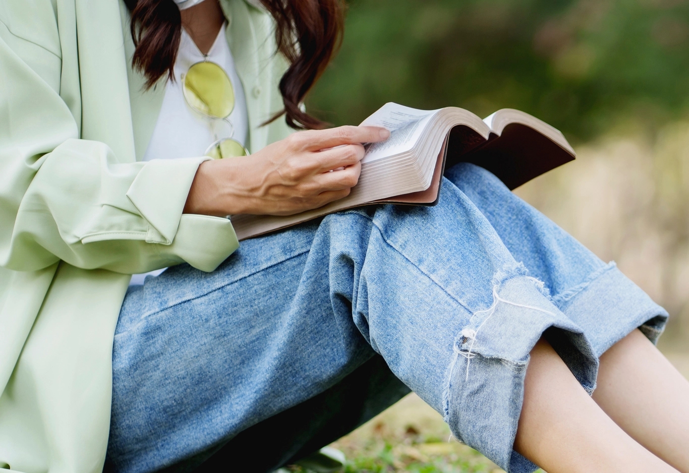 Person som sitter utomhus i jeans och en lätt jacka och läser en bok med solljus på sidorna.
