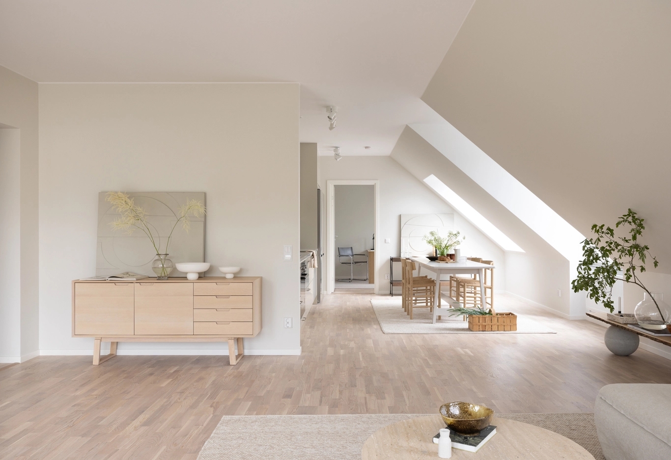 Ljust, minimalistiskt vardagsrum och matplats med ljusa trämöbler, vita väggar och två stora snedställda fönster.