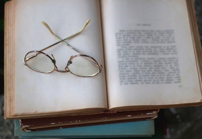 Ett par glasögon vilar på en öppen bok med suddig text, ovanpå en trave böcker.