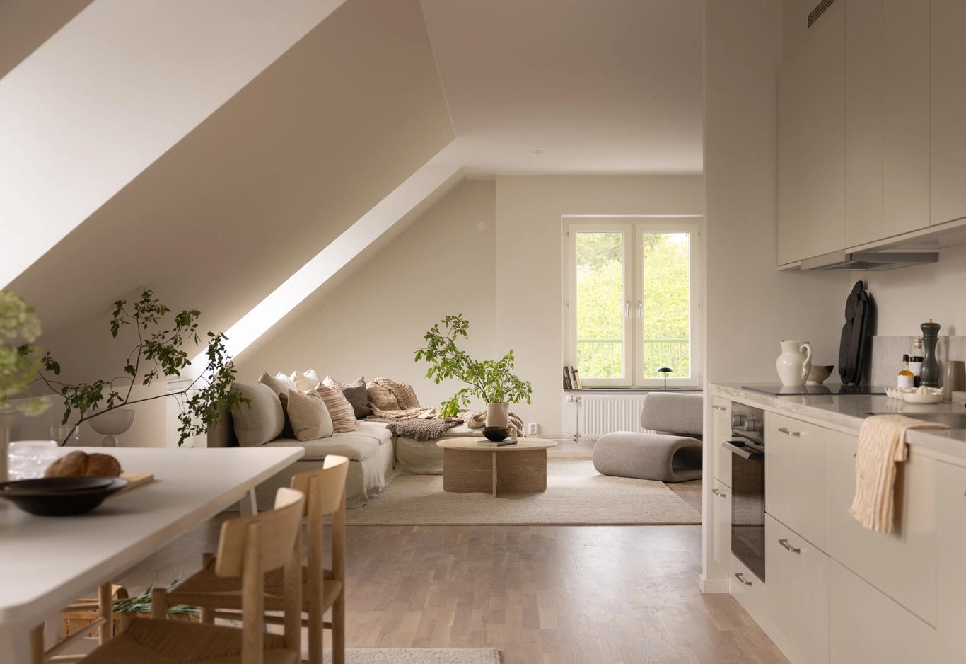 Modern vindslägenhet med neutral inredning. Kök, matbord och en mysig vardagsrumsdel med växter vid fönstret.