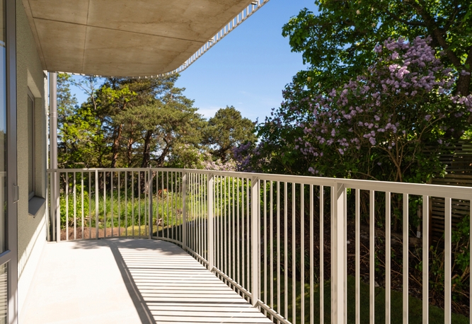 Stor balkong med vitt metallräcke och utsikt mot gröna träd och lila syrener under en solig blå himmel.
