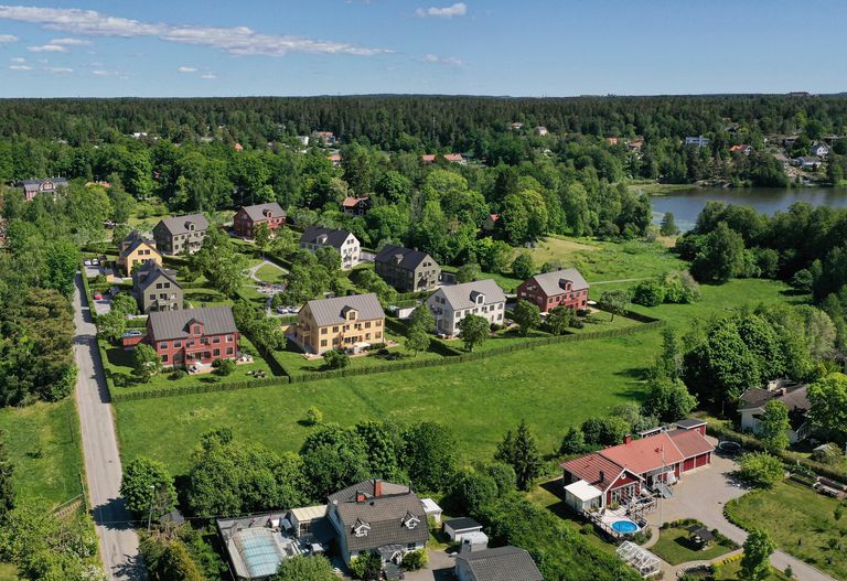 Helikoptervy över bostadsområde med flera hus omgivna av grönska, en väg och en närliggande sjö under en klarblå himmel.