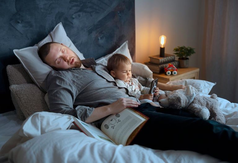 En man sover på en säng med ett litet barn som sitter bredvid honom och håller i en smartphone. Barnet är omgivet av mjuka leksaker och en bok ligger öppen bredvid dem. En liten lampa står på nattduksbordet.