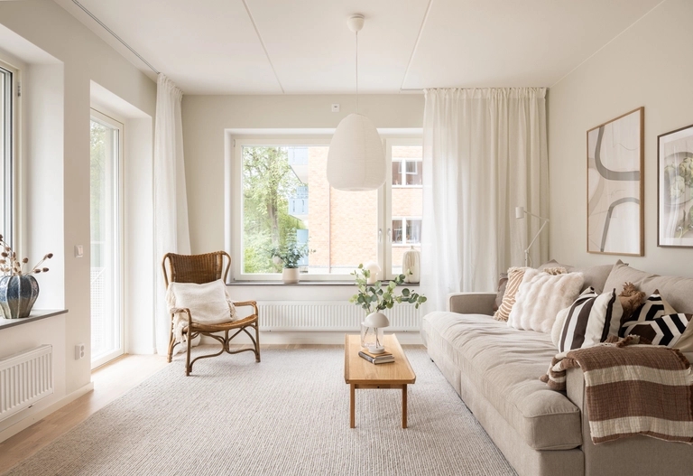 Ljust vardagsrum med en beige soffa, soffbord i trä, korgstol, stora fönster och neutral inredning.
