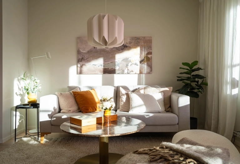 Soligt vardagsrum med en vit soffa, orange kuddar, ett runt soffbord och väggkonst ovanför soffan.