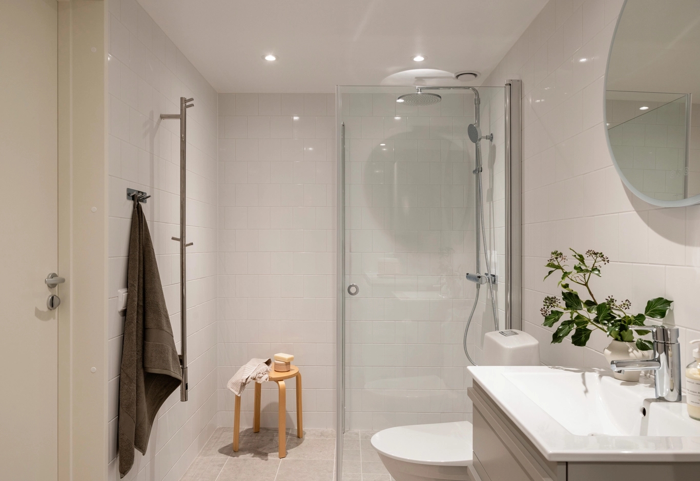 Modernt badrum med vita väggar, dusch i glas, rund spegel, handfat och en liten träpall med handduk och toalettartiklar.