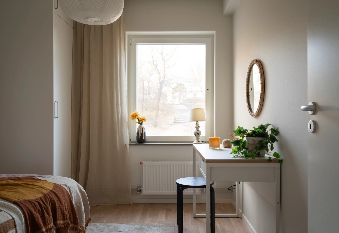 Ett litet, prydligt sovrum med säng, fönster, skrivbord, pall, lampa och växter i naturligt ljus.