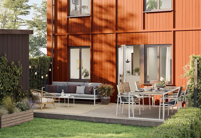 En uteplats med röd exteriör i trä har ett matbord, stolar och en soffa. Det finns växter och träd runt omkring.