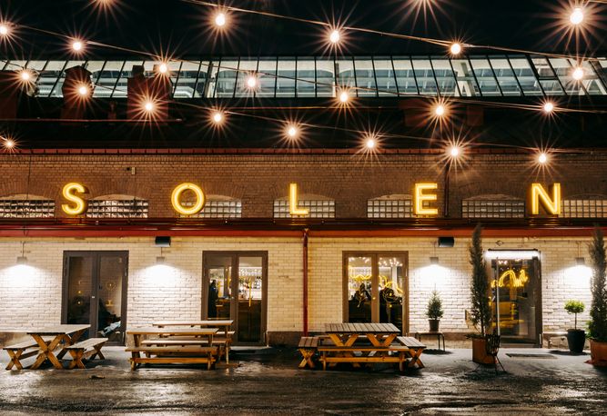 Uthomhusvy av Restaurang Solens tegelbyggnad med "SOLEN" i upplysta bokstäver. Uteservering med ljusslingor framför. Kvällsljus.