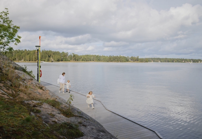 Tre personer går längs en träbrygga vid ett lugnt vatten, med träd och klippor i bakgrunden under en molnig himmel.