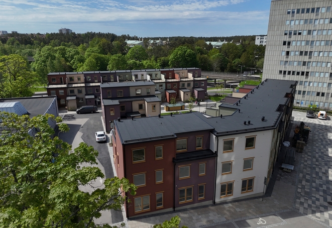 Moderna radhus i varierande färger och en hög lägenhetsbyggnad i ett lummigt bostadsområde under en blå himmel.