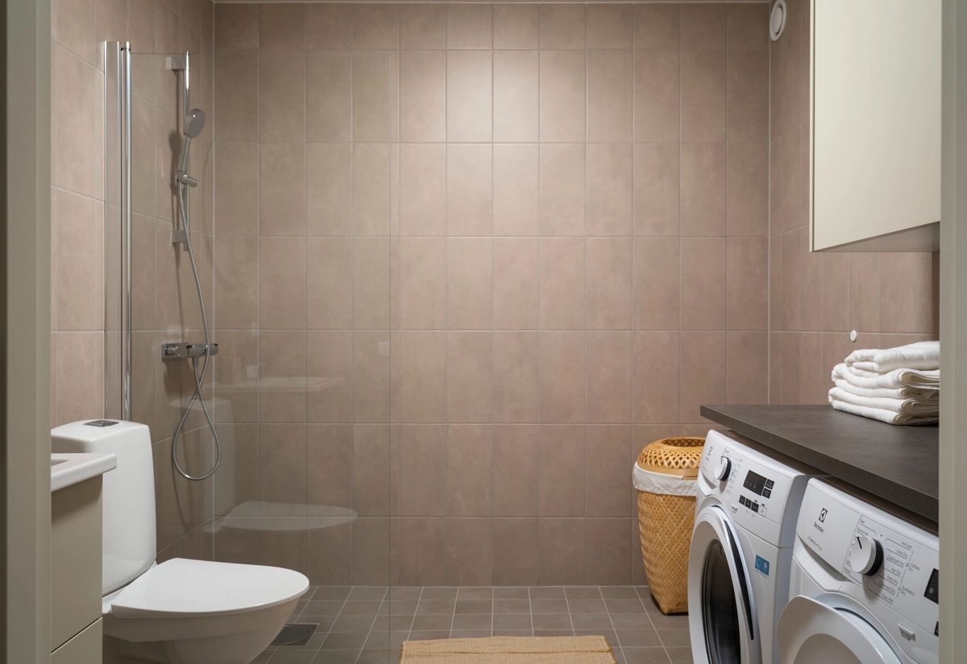 Ett badrum med toalett, dusch, tvättmaskin, torktumlare, tvättkorg och handdukar på en bänk.