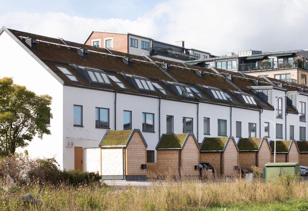 Vita radhus med mörka tak, takfönster och små trähus med spetsiga tak framför. I förgrunden gräs, i bakgrunden flerbostadshus.