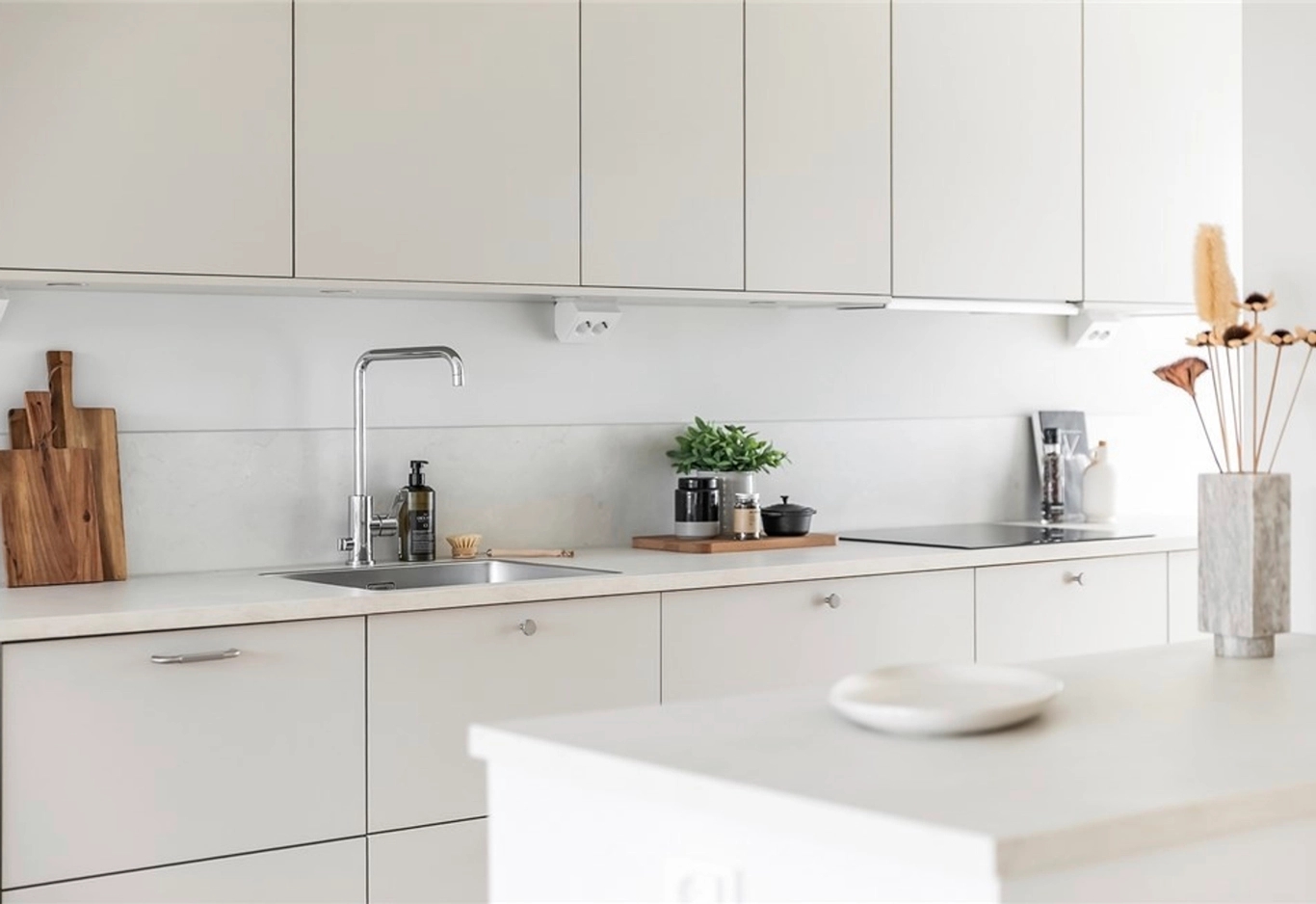Ett modernt kök med vita skåp, diskbänk, krukväxter, skärbrädor och minimal bänkinredning.