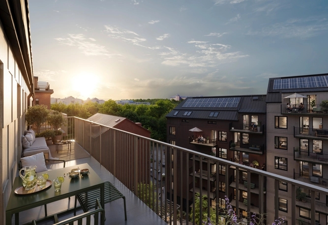 Bred balkong med sittplatser och bord med utsikt över moderna flerbostadshus och grönska vid solnedgången.
