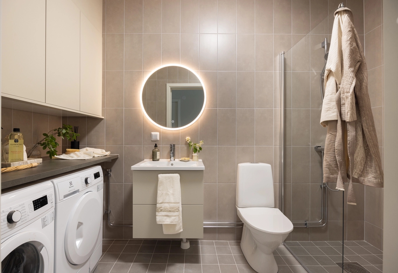 Modernt badrum med tvättmaskin, rund spegel med bakgrundsbelysning, handfat, toalett och hängande badrockar bredvid en dusch med duschväggar.