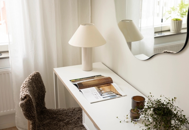 Ett vitt skrivbord med en uppslagen tidning, en lampa, en liten växt och två stearinljus står vid ett fönster med en vågig spegel ovanför.