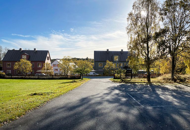 Vy över en asfalterad väg som leder mot två hus, träd och en klarblå himmel. Solljus kastar skuggor på vägen och gräsmattan, med inslag av höstlöv.