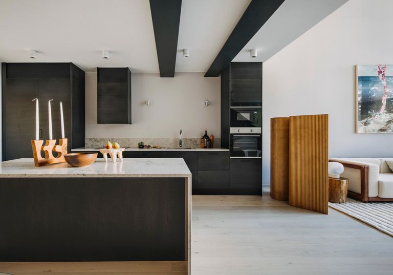 En möblerad matplats i anslutning till ett arbetskök med litens köksö syns i förgrunden. I bakgrunden stora fönsterpartier med utblick mot trädgård.