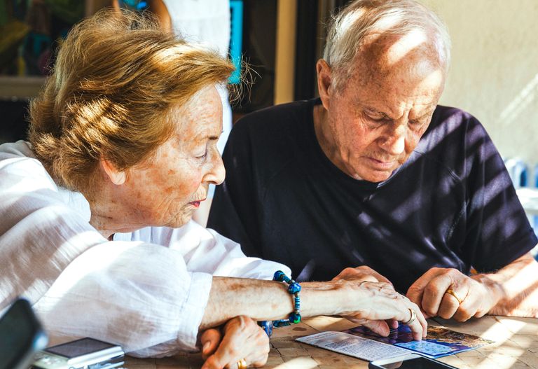 Två äldre personer sitter vid ett bord och granskar noga papper eller dokument i starkt naturligt ljus.