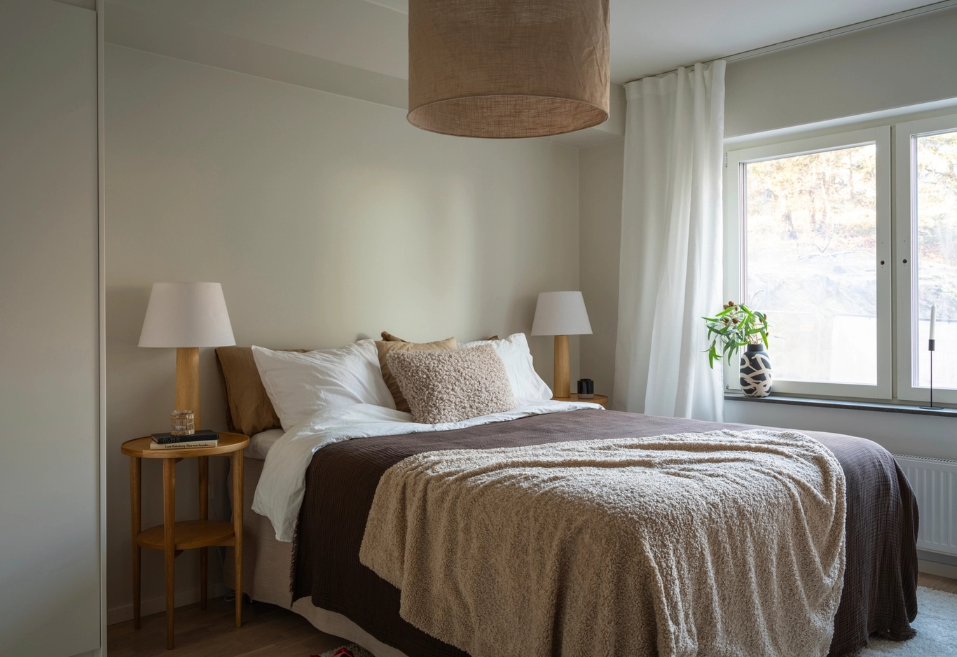 En prydligt bäddad säng med beige och bruna sängkläder, två sidobord med lampor och ett fönster med vita gardiner.
