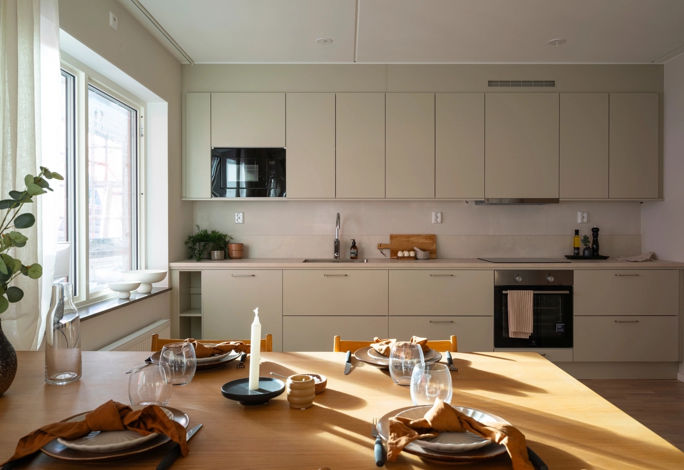 Modernt kök med beige skåp, inbyggd ugn och ett matbord med plats för fyra personer vid ett stort soligt fönster.