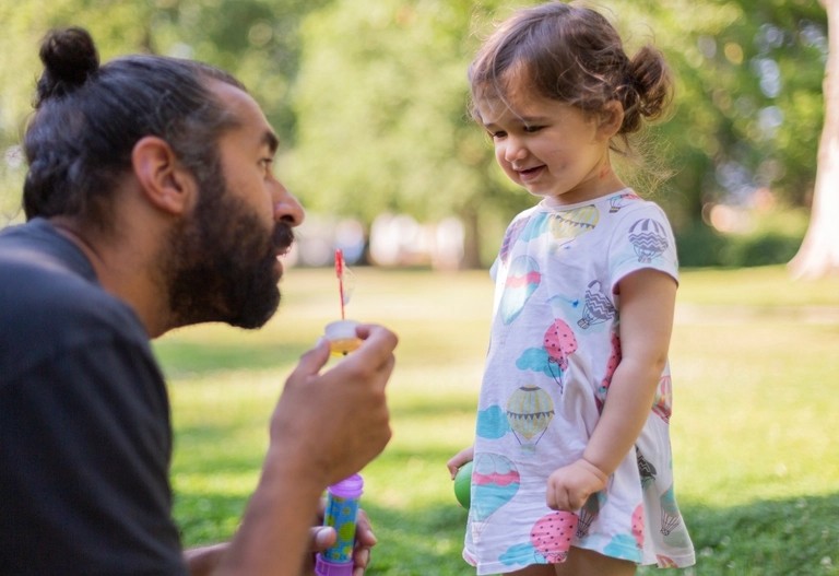 En vuxen förbereder sig på att blåsa bubblor medan en ung flicka i en mönstrad klänning ler utomhus i en park.