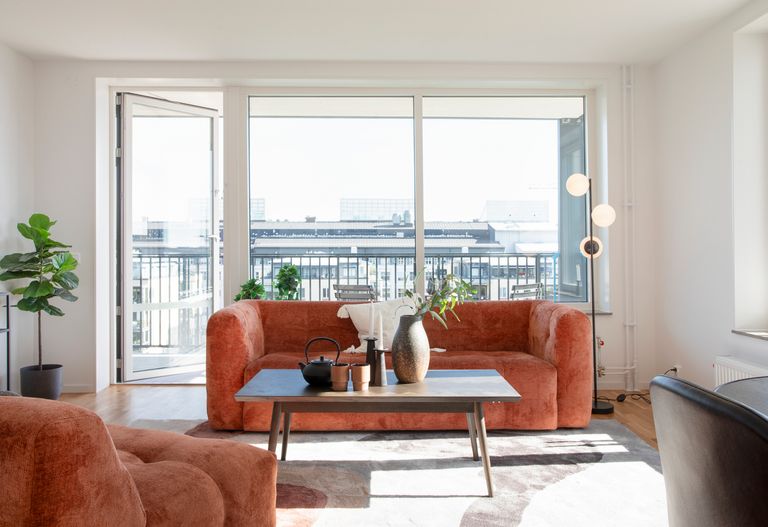 Ljust vardagsrum med ett stort fönster, orange soffa, soffbord och utsikt över balkongen, dekorerat med växter