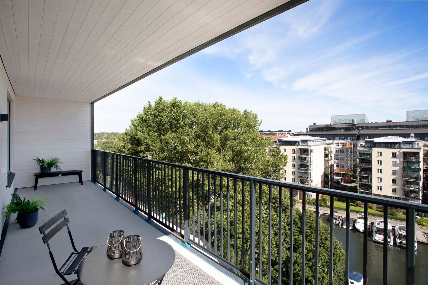Solig urban balkong med utsikt över byggnader och träd, möblerad med bänk och krukväxter.